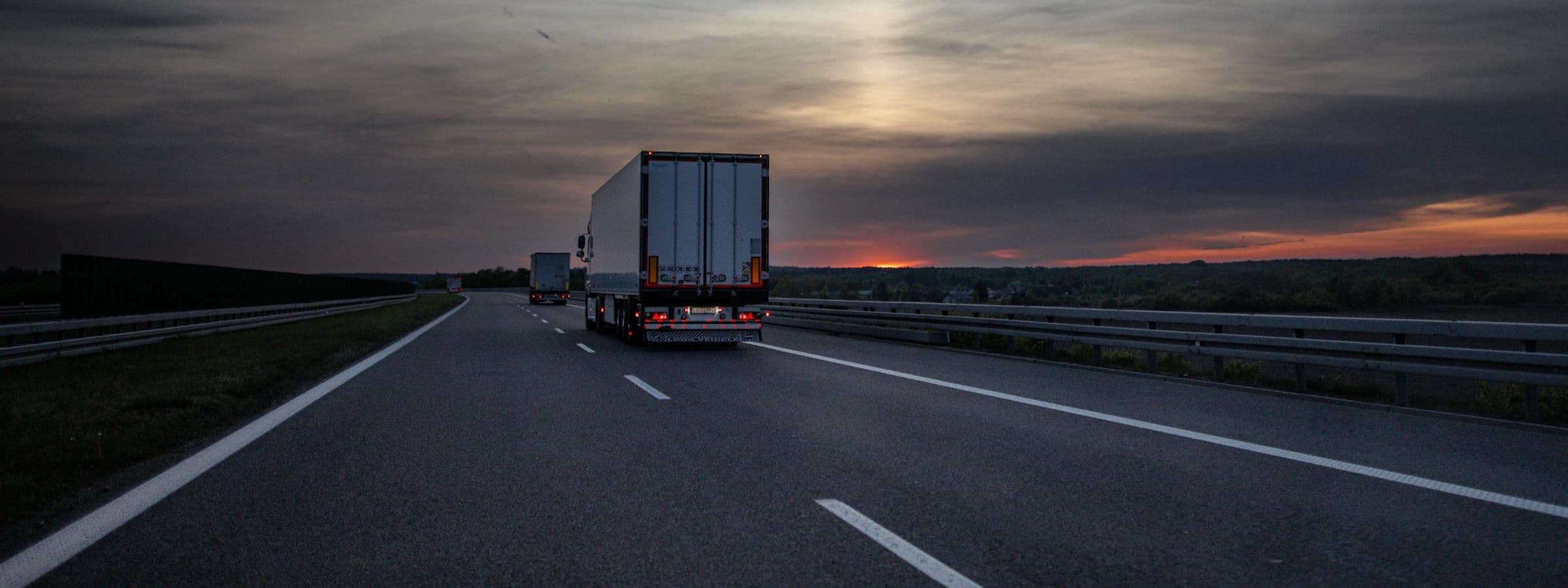 A tractor-trailer travelling an open highway at dusk, with lane markings running toward a low sun on the horizon.